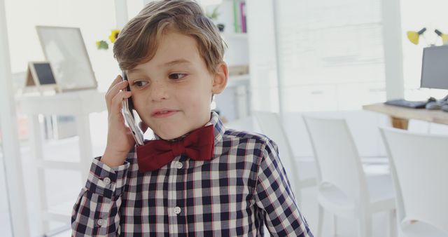 Young boy wearing plaid shirt and red bow tie talking on smartphone inside office. Use for themes of youth communication, tech-savvy kids, business, education, or professional environments.