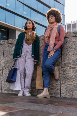 Diverse young women leaning on urban wall holding shopping bags with backpacks and scarves