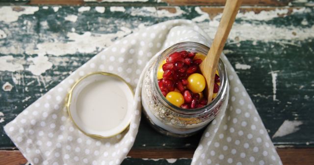 Shows a glass jar filled with layers of yogurt, pomegranate seeds, and yellow berries, placed on a rustic wooden table with a polka dot cloth. Ideal for use in health, wellness, food, recipe blogs, or nutrition publications. Can also be relevant for promotions of healthy eating habits, breakfast ideas, or rustic culinary themes.