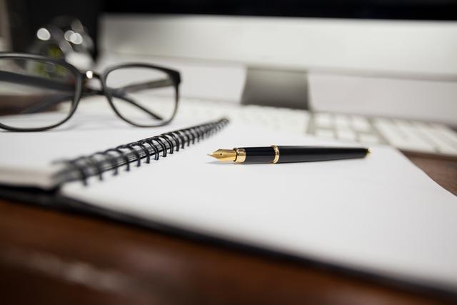 Close-up of a spiral notepad with a fountain pen and reading glasses on a wooden desk. Ideal for illustrating concepts related to office work, studying, planning, writing, and professional environments. Suitable for use in articles, blogs, and websites about productivity, business, education, and office supplies.