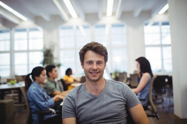 Portrait of man smiling at camera while colleagues working in background at the office