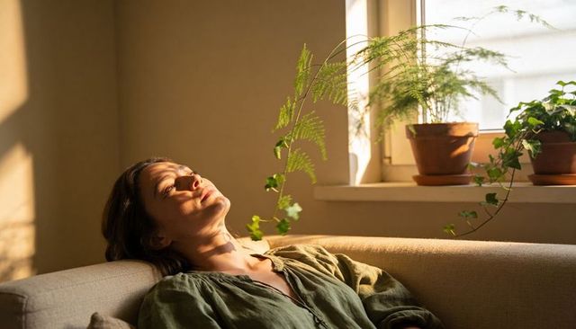 Serene woman relaxing on sofa, basking in warm sunlight with potted ferns on windowsill