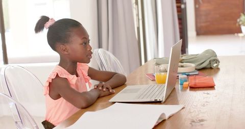 Young girl studying at home using laptop on dining table with school supplies