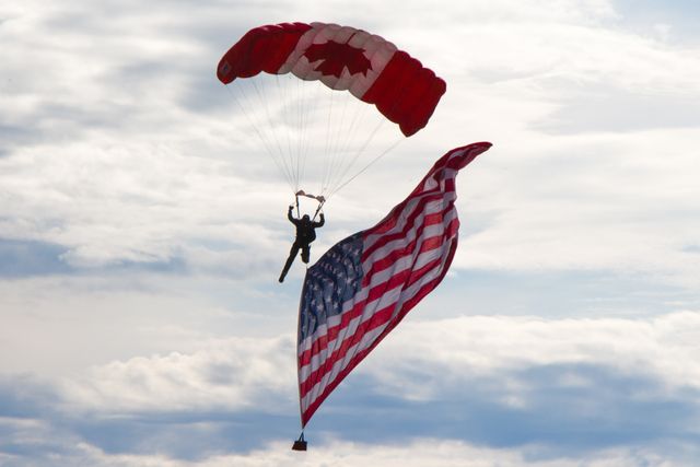 A skydiver is parachuting with a Canadian flag and holding an American flag against a cloudy sky. This image captures the spirit of adventure and freedom while highlighting the camaraderie between Canada and the USA. Perfect for topics on international relations, patriotic events, adrenaline sports, and freedom or unity-themed promotions.
