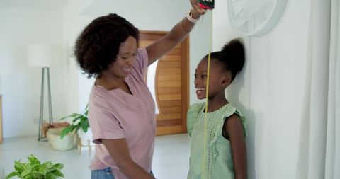 Mother measuring daughter's height with tape measure while smiling in bright modern foyer
