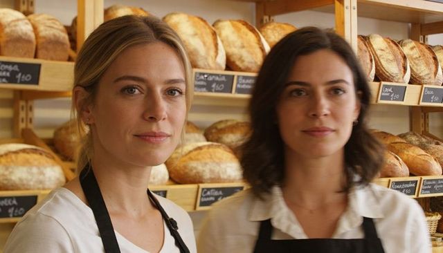 Two Bakers Standing in Artisan Bakery Surrounded by Rustic Sourdough Loaves on Wood Shelves
