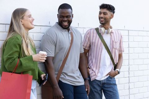 Diverse young friends laughing and leaning on white brick wall with coffee and shopping