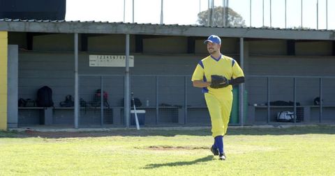 Middle-aged baseball player walking on sunlit field holding glove by dugout, smiling