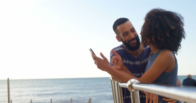 Romantic diverse couple standing by railing, talking and using smartphone on sunny beach, copy space. Summer, vacation, communication, romance, love, relationship, free time and lifestyle, unaltered.