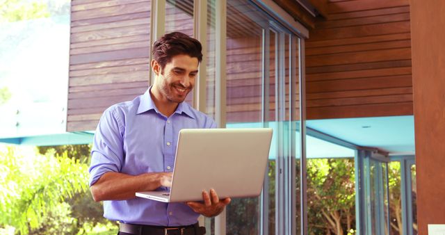 Professional man in blue shirt happily working on laptop at outdoor office with greenery in background. Perfect for illustrating remote work, modern business environments, freelance lifestyle, digital nomad trends, and technology in professional settings.