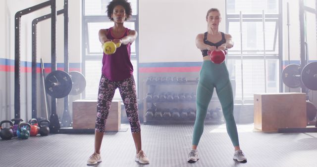 Image of two diverse women working out at a gym doing kettlebell swings in unison. Exercise, fitness and healthy lifestyle.