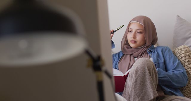This image features a young Muslim woman wearing a hijab, sitting indoors, studying with a notebook and pen. She is concentrating on her work, creating a thoughtful atmosphere. Ideal for educational content, blog posts about studying habits, remote learning, or articles promoting women's education in diverse cultures. Also useful for visual materials focusing on inclusion, diversity, and lifestyle concepts.