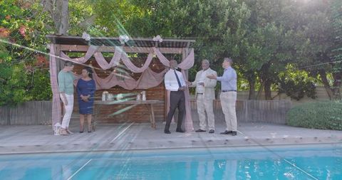Friends chatting by pool with pink pergola drapery during sunlit backyard celebration