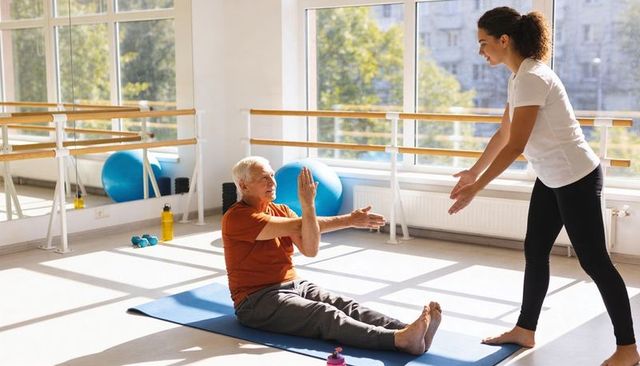 Senior man practicing assisted balance exercise with female trainer in bright fitness studio