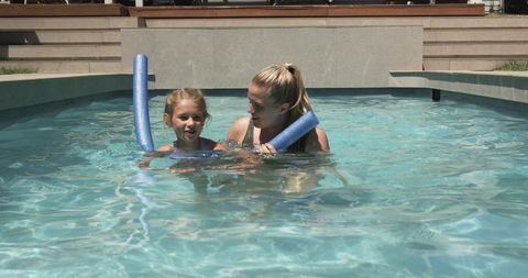 Female guardian teaching young girl to swim with blue pool noodles in sunlit backyard pool