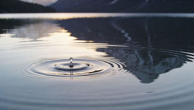 Water droplet creating concentric ripples on glassy alpine lake reflecting mountain at sunset