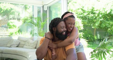 African american couple laughing and hugging in sunlit living room overlooking garden