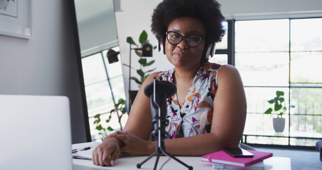 Black woman confidently recording a podcast in her home office, with microphone and headphones. Ideal for content related to entrepreneurship, remote work, podcasting, modern work environment, or personal branding.