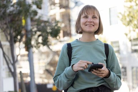 Young woman smiling while using smartphone on sunlit city sidewalk with backpack