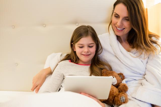 Image shows a mother and daughter sitting on a bed and using a digital tablet together. The mother is wearing a white robe, and the daughter is holding a teddy bear. Both are smiling and enjoying their time together, showcasing family bonding and technology use in a cozy home environment. This image can be used for themes related to family, home life, parenting, technology, and relaxation.