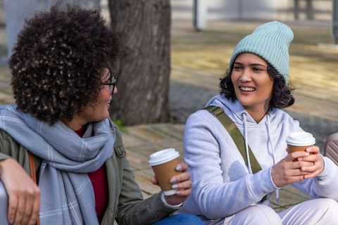 Urban friends sharing coffee and conversation on plaza bench wearing beanie and scarf