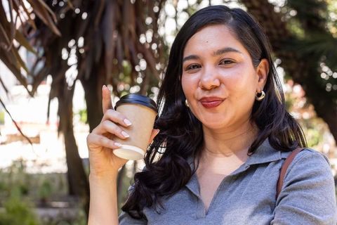 Smiling woman holding takeaway coffee cup in sunlit urban park portrait casual lifestyle
