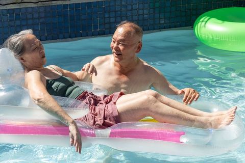 Senior asian couple relaxing on clear inflatable lounger floating in sunny backyard pool