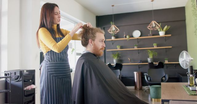 A hairdresser is styling the hair of a man in his midthirties at a modern salon. The hairdresser wears striped apron, and the man is seated, covered with a cape. The salon features potted plants and hanging light fixtures, creating a relaxed and contemporary atmosphere. Ideal for ads related to grooming, professional hair care services, modern barbershops, or cosmetic brands.