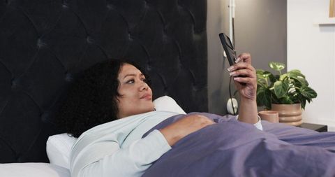 African American woman reclining in bed holding smartphone, browsing and messaging