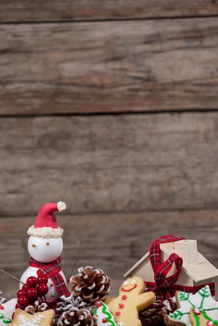 Festive snowman surrounded by Christmas decorations including pine cones, gingerbread cookies, and red berries. Wooden background adds a rustic touch. Ideal for holiday greeting cards, festive advertisements, and seasonal social media posts.