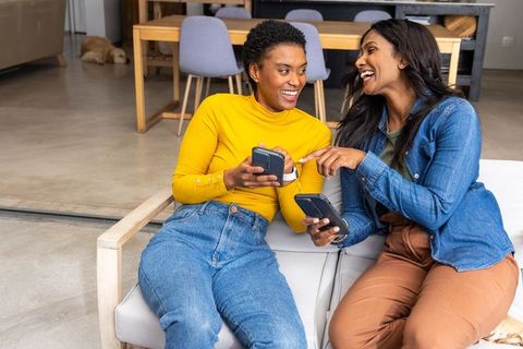 Diverse female friends laughing and sharing phones while relaxing on modern living room loveseat