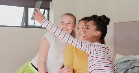 Diverse friends smiling while taking group selfie on bed bathed in natural light