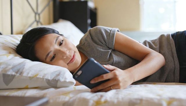 Reclining asian woman using smartphone on bed, casual grey tee, relaxed daylight moment