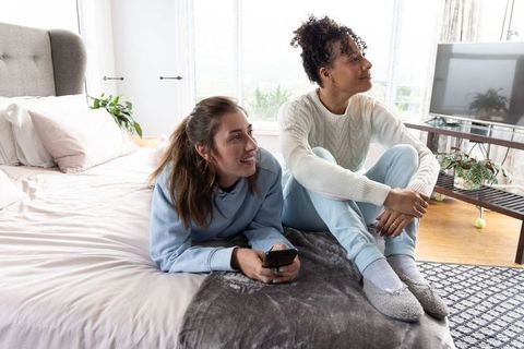 Two women relaxing on bed by bright windows, cozy morning bonding with smartphone