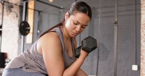 Asian woman performing single-arm dumbbell curl on bench in gym focusing on strength