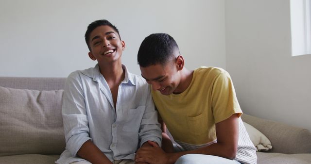 Happy biracial gay male couple sitting on sofa laughing. staying at home in isolation during quarantine lockdown.