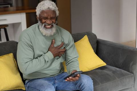 Senior african american man clutching chest while holding phone sitting on sofa in pain