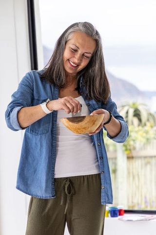 Smiling mature woman mixing ingredients in wooden bowl near sliding glass door