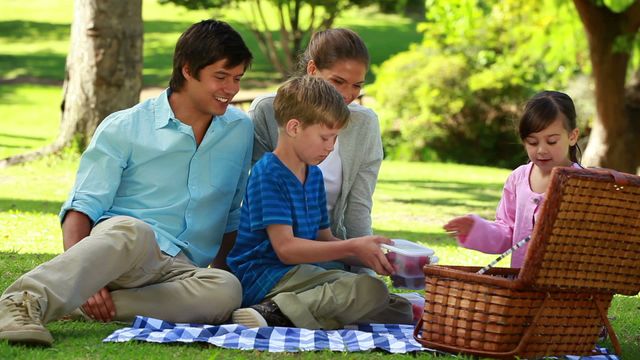 A family with children is having a picnic in a lush green park, sitting on a blanket and likely enjoying quality time together. Their joyful expressions add warmth and an essence of togetherness to the scene. Ideal for use in websites about family activities, leisure content, or promoting parks and outdoor events. This scene beautifully captures the essence of outdoor family bonding and relaxation.