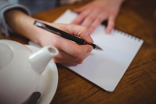 Woman writing in a notepad at a cafe table, ideal for content about productivity, journaling, and creative processes. Perfect for blogs, articles, and advertisements focused on coffee shop environments, casual work settings, and lifestyle imagery.
