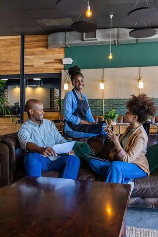 African american server smiling and chatting with customers on leather sofa in cozy cafe