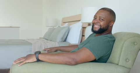 Smiling man relaxing in green armchair in cozy neutral bedroom with watch and headboard