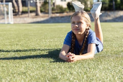 Young girl lying on sunlit soccer field wearing braids and white sneakers