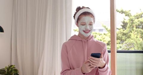 Smiling woman wearing facial mask and headband, using smartphone during calm self-care routine