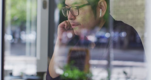 Man wearing wireless earbuds, deeply focused, working inside a café. Scene shot through a window, showing reflection and urban greenery outside. Perfect for illustrating concepts of remote work, technology use, concentration, and modern professional lifestyle. Suitable for business, technology, and lifestyle content.