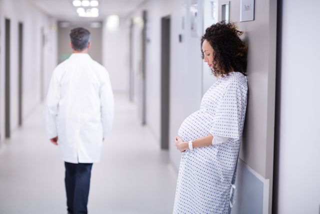 This image shows a pregnant woman standing in a hospital corridor, leaning against the wall, with a doctor walking away in the background. It can be used for articles or advertisements related to maternity care, prenatal health, hospital services, and healthcare facilities. It is also suitable for educational materials on pregnancy and medical care.