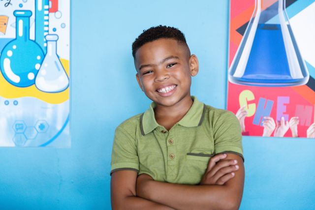 This image depicts a cheerful African American elementary schoolboy standing with his arms crossed in front of colorful science posters. Ideal for educational materials, school brochures, STEM program promotions, and websites focusing on children's education and learning environments.