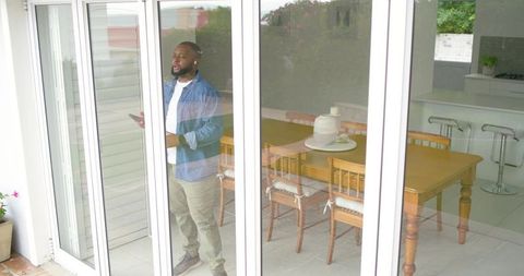 African american man standing behind sliding glass doors holding phone by dining table