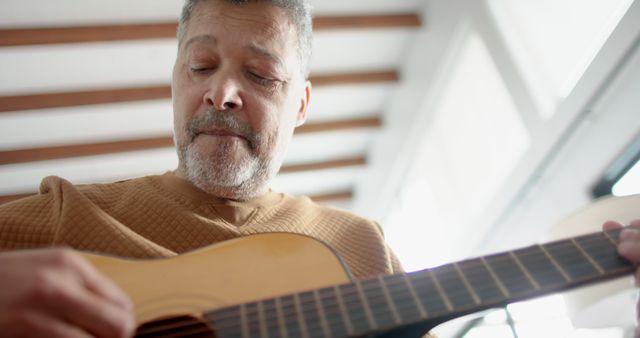 Senior man playing acoustic guitar in bright, sunlit room, expressing enjoyment and relaxation. Perfect for themes on music, hobbies, senior living, and home activities. Excellent for promoting music lessons, retirement lifestyle, and leisurely pastimes.