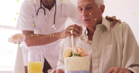 Senior man blowing candle on cake with caregiver offering gentle support during birthday